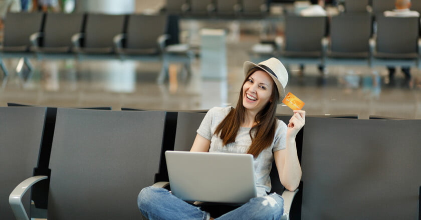 Young smiling traveler tourist woman in hat sit with crossed legs, working on laptop hold credit card wait in lobby hall at airport. Passenger traveling abroad on weekends getaway. Air flight concept.