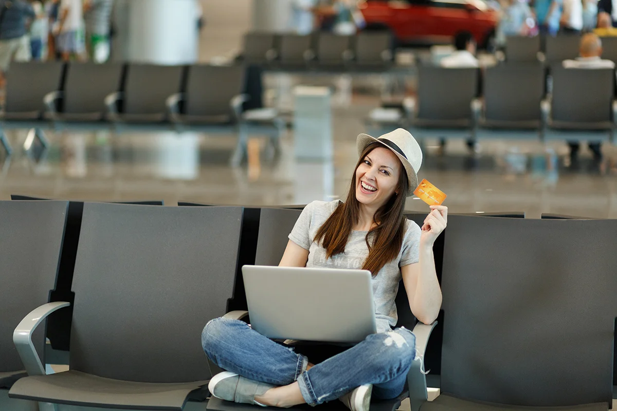 Young smiling traveler tourist woman in hat sit with crossed legs, working on laptop hold credit card wait in lobby hall at airport. Passenger traveling abroad on weekends getaway. Air flight concept.